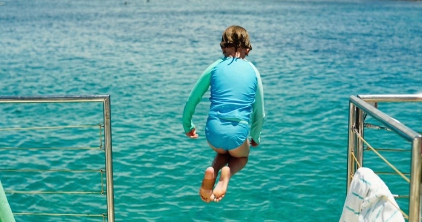 Child in a blue rash guard jumping off the back of a boat into turquoise water