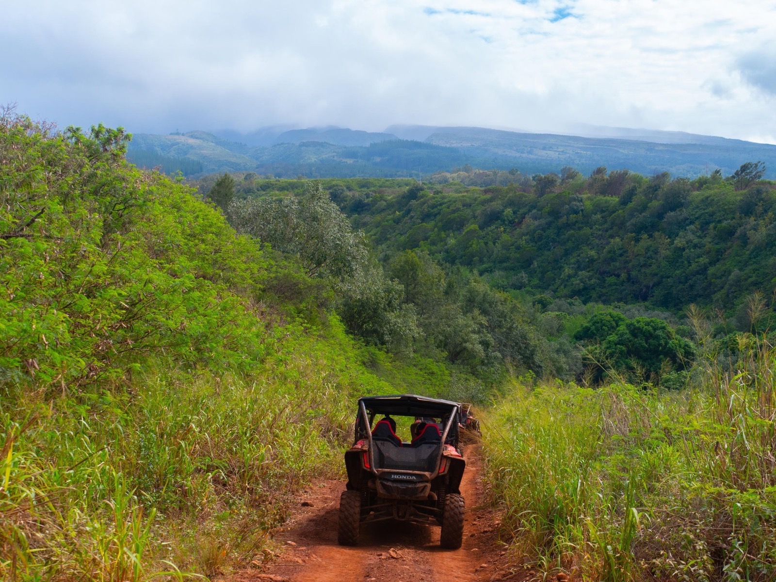 A Honda side-by-side drives down a red dirt trail toward a green valley with cloud-covered West Maui mountains behind