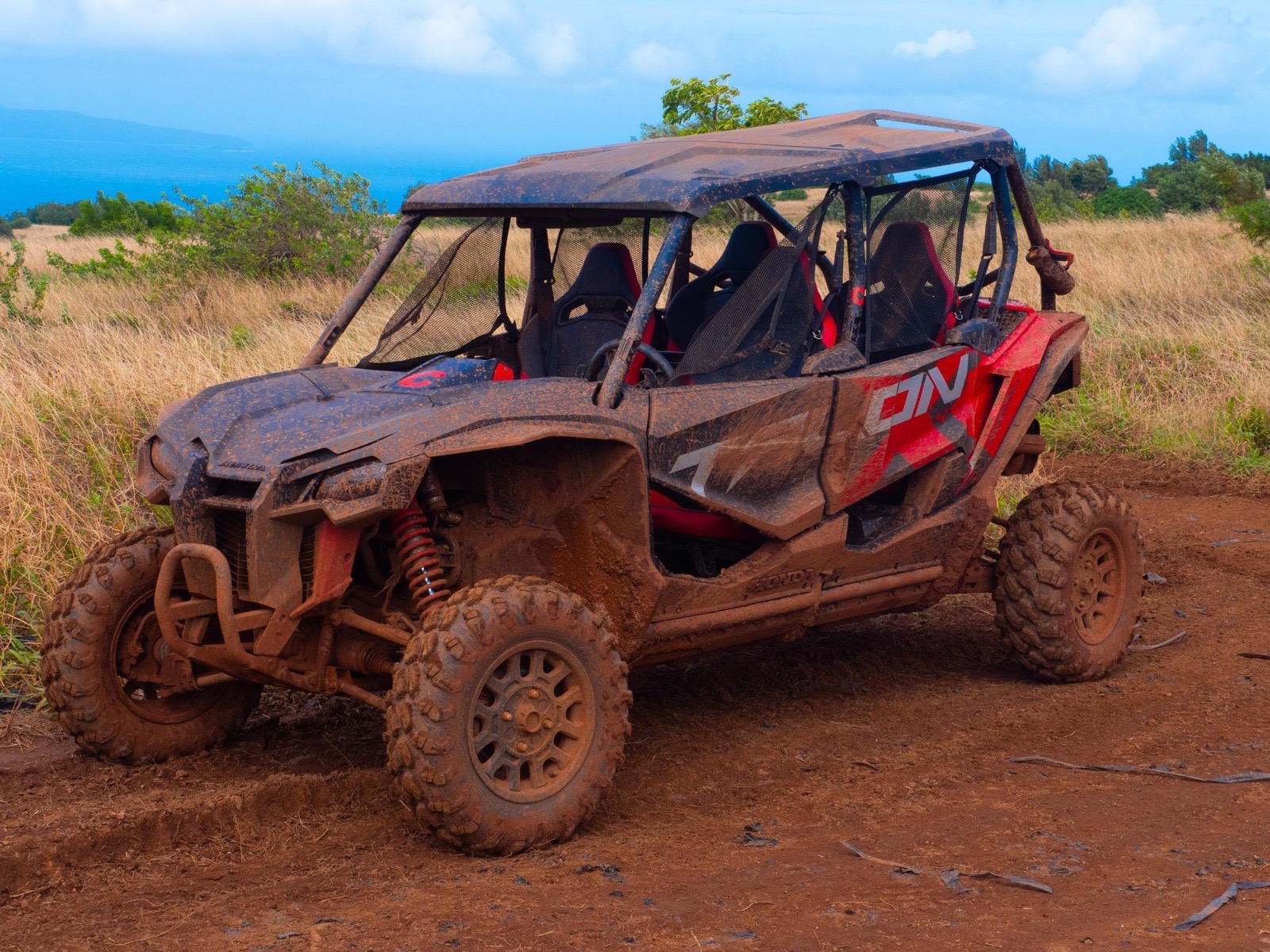 A muddy red Honda four-seat side-by-side parked on red dirt beside golden grass, with the ocean visible beyond