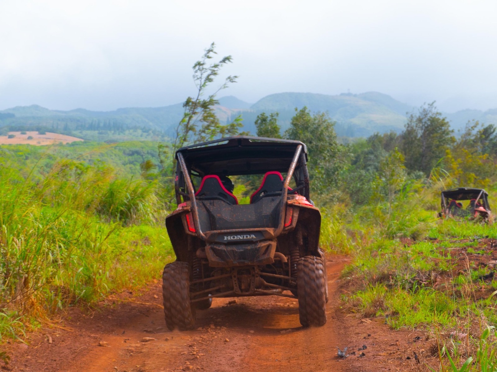The back of a Honda side-by-side on a red dirt ridge trail with misty hills and a second vehicle behind