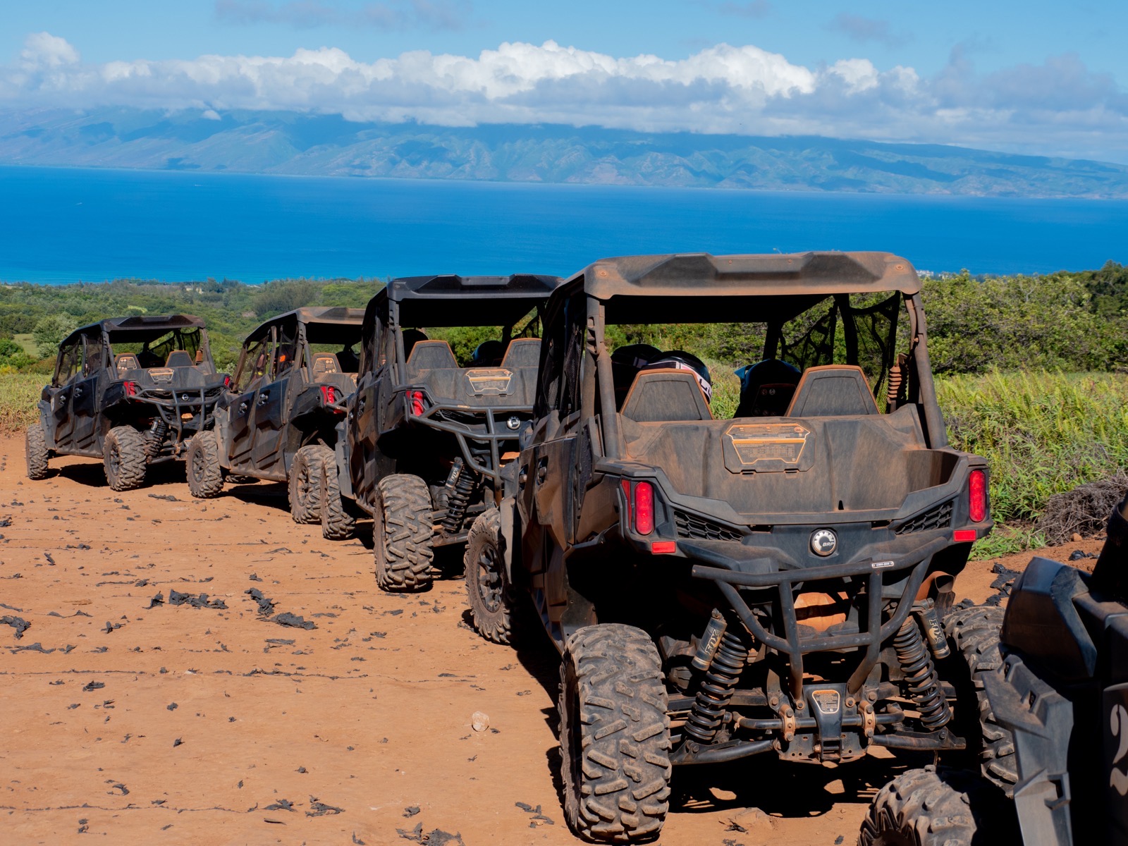 A line of four Can-Am side-by-sides parked on a red dirt overlook above a deep blue ocean with another island on the horizon