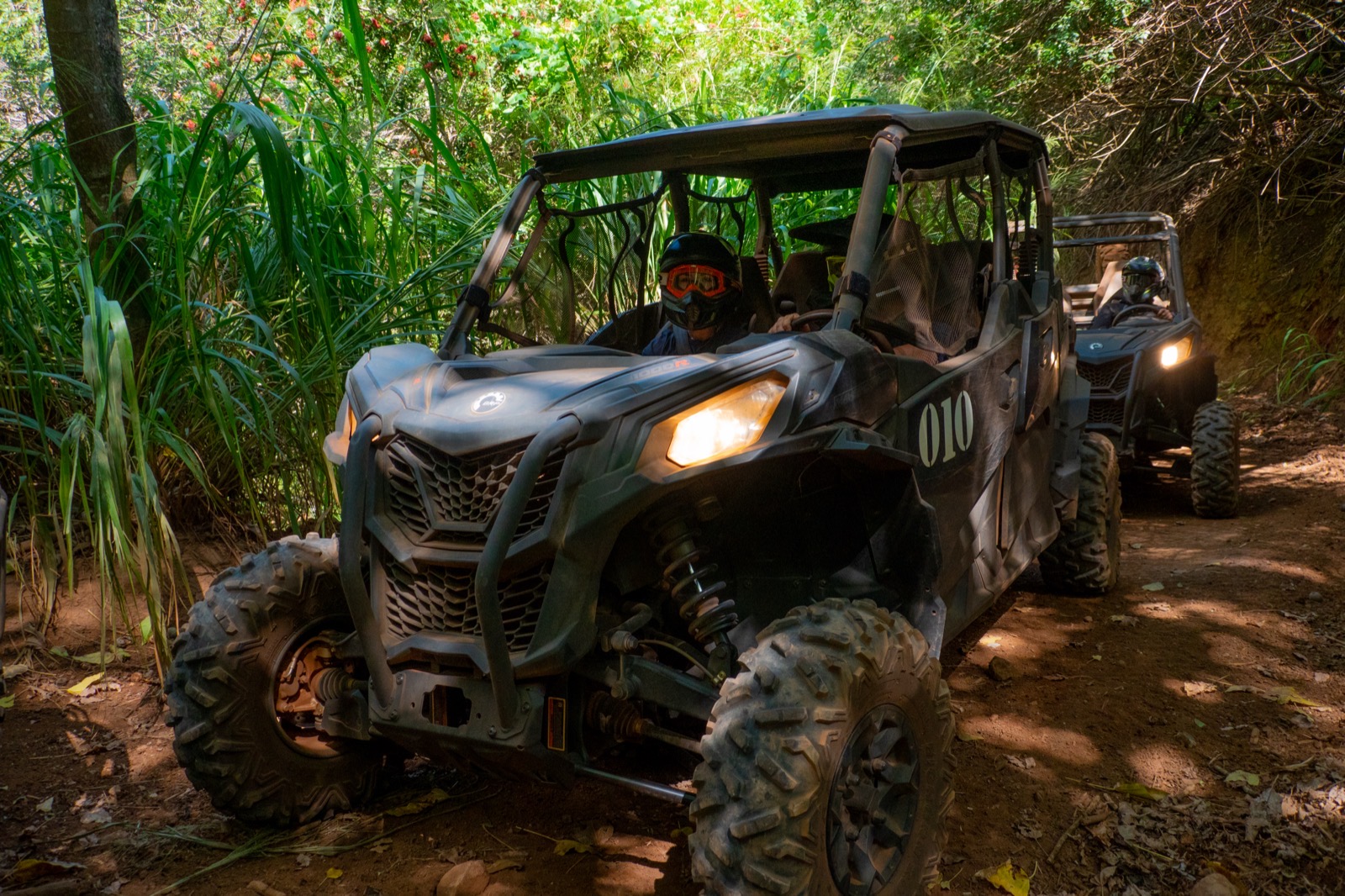 Two Can-Am side-by-sides with headlights on drive single file through a shady bamboo and jungle trail