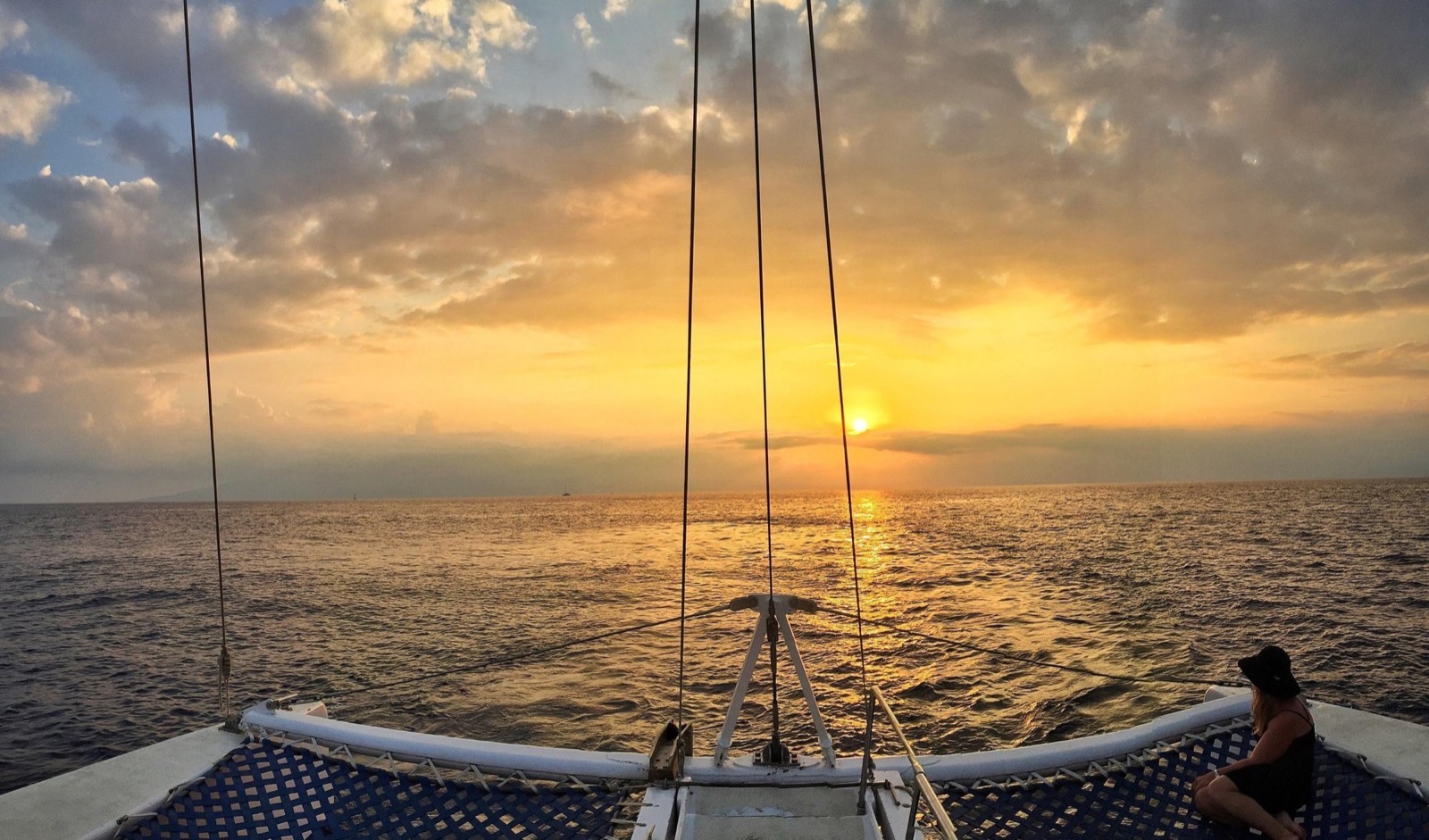 View from the bow of the catamaran toward a low orange sun, with a guest in a black hat sitting on the net