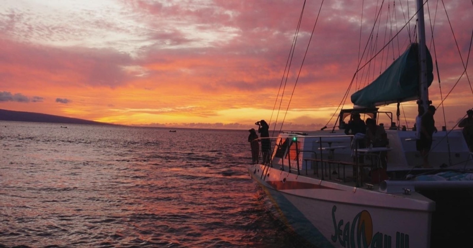 Silhouetted guests at the rail of a Sea Maui catamaran against a fiery red and orange sunset