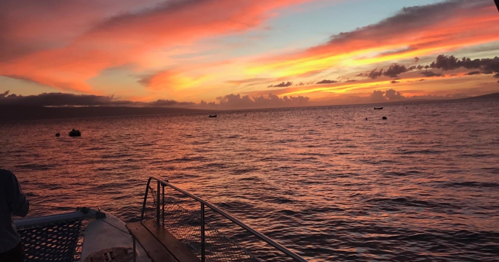 Orange and pink sunset sky over the ocean seen from the bow of the catamaran, with the dark shape of Lanai on the horizon