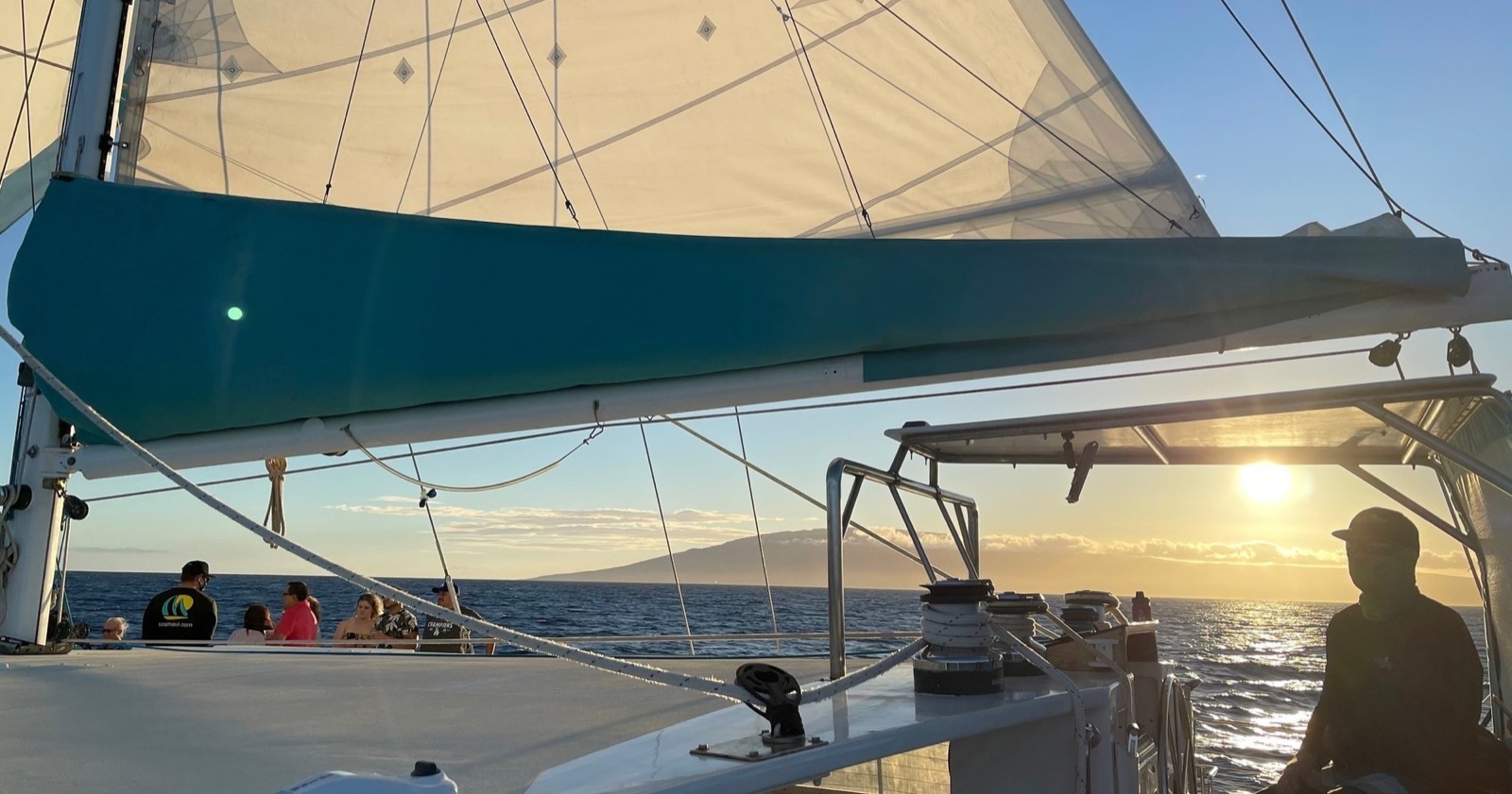 Guests standing on the deck under the teal-covered sail as the sun sinks toward the mountains of Lanai