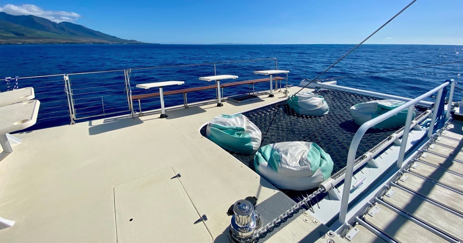 Bean bag chairs on the bow trampoline and rail-side tables on deck, with the West Maui mountains in the distance