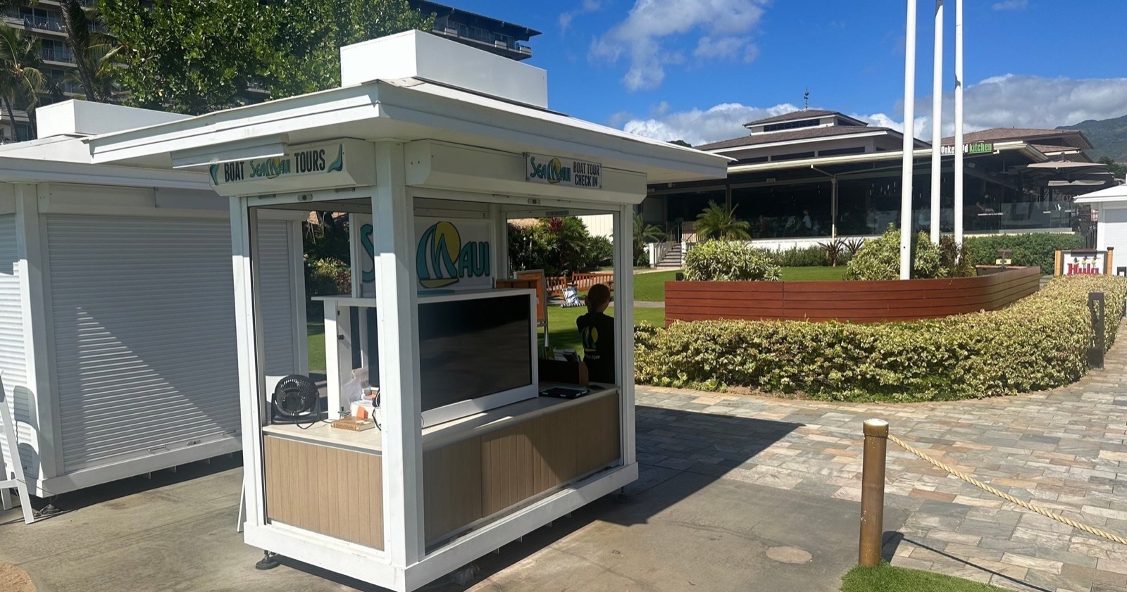 The white Sea Maui boat tour check-in kiosk on the Whalers Village beach walk, with Hula Grill in the background