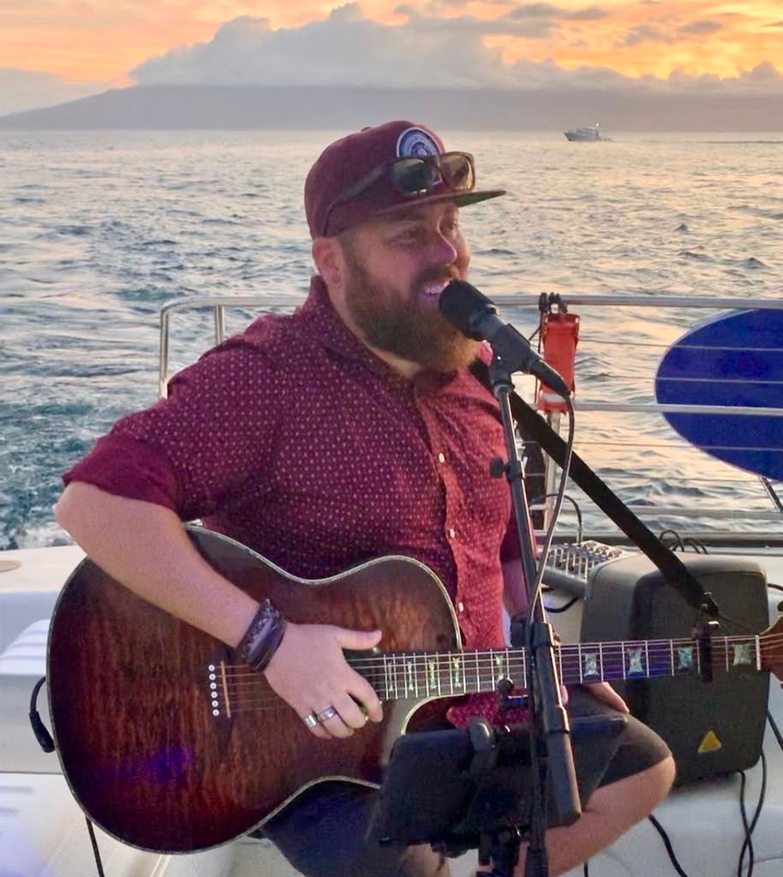 A musician singing and playing acoustic guitar on the back of the boat with a pastel sunset over the ocean behind him