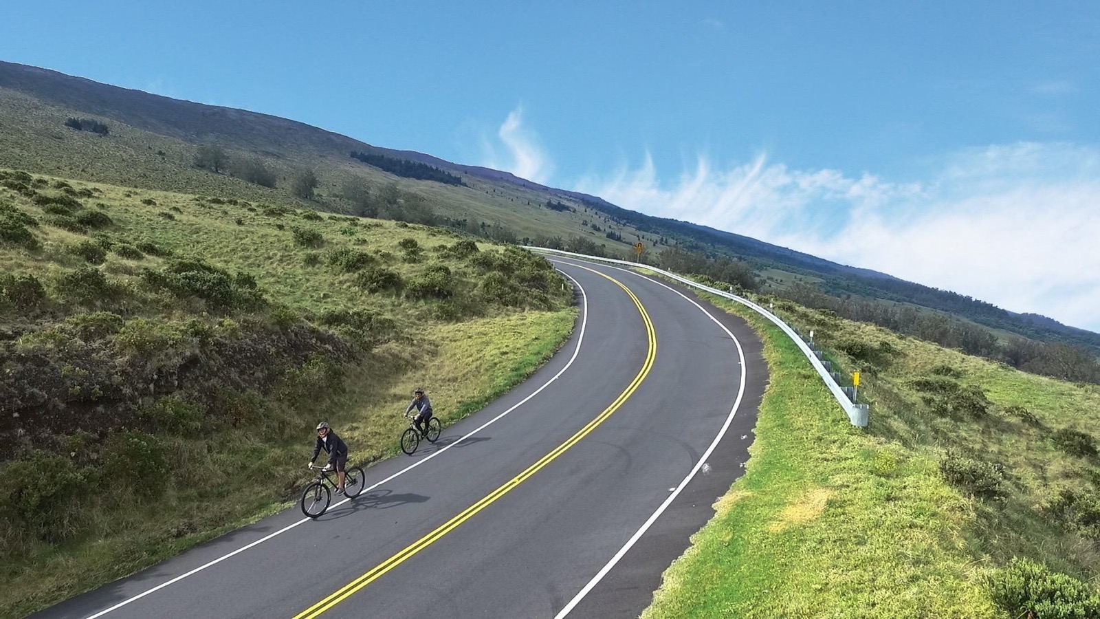 Two cyclists in helmets ride a curving two-lane road on the upper slopes of Haleakala under a blue sky with wispy clouds