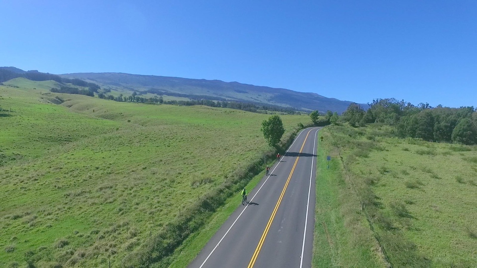 Three riders coast down a straight paved road through open green pasture with the long slope of Haleakala behind them