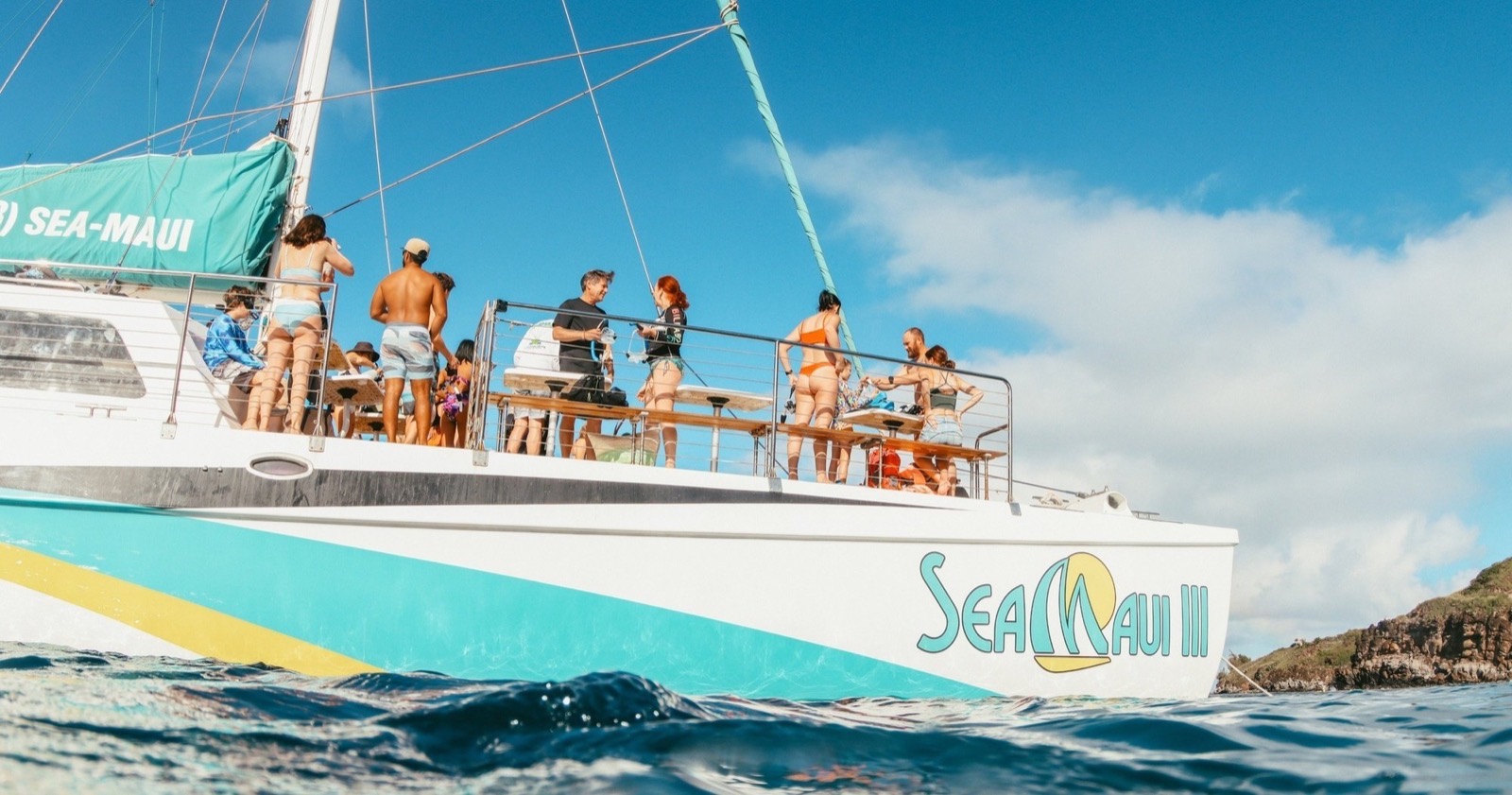 Guests gathered on the open deck of the Sea Maui III catamaran while anchored near a rocky Maui shoreline