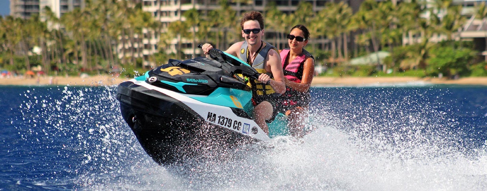A driver and passenger in sunglasses and life jackets kicking up spray on a Sea-Doo with the palm trees and hotels of Kaanapali Beach behind them