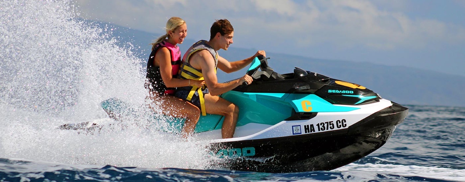 A smiling couple leaning into a turn on a teal Sea-Doo, with a wall of white spray behind them and the hazy outline of Lanai in the distance