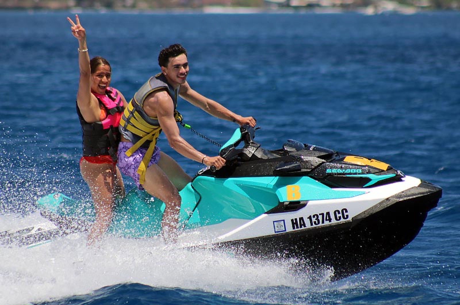 A passenger raising a peace sign behind a driver on a Sea-Doo, the engine cut-off lanyard visible on his wrist, over bright blue open water