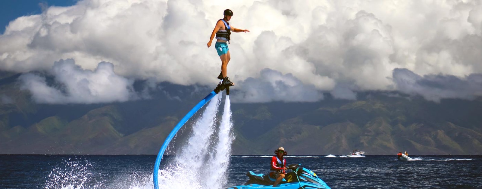 A flyboarder standing high above the sea on twin jets of water, with the green West Maui mountains and a jet ski below him