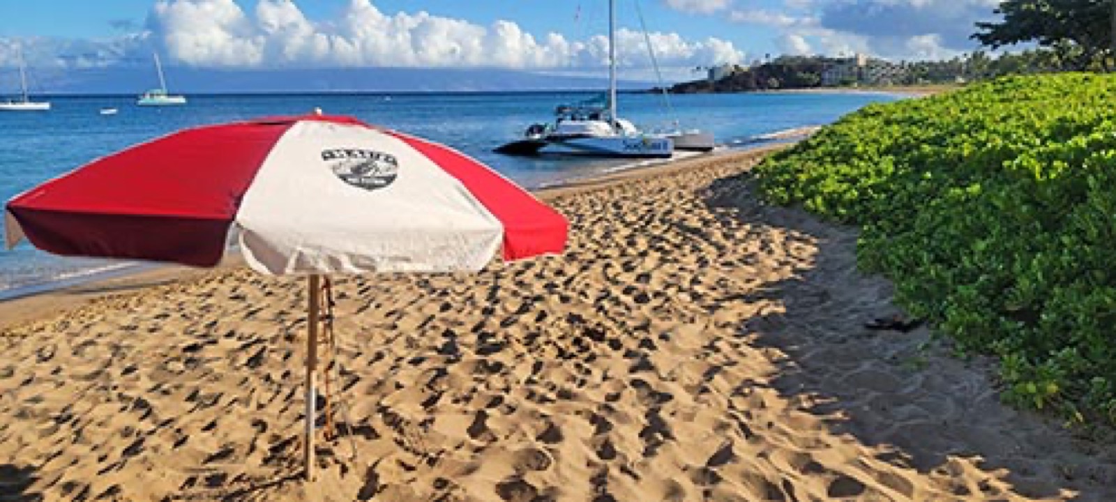 A red and white beach umbrella planted in the sand at Kaanapali Beach, with a catamaran at the water's edge and the shoreline curving away