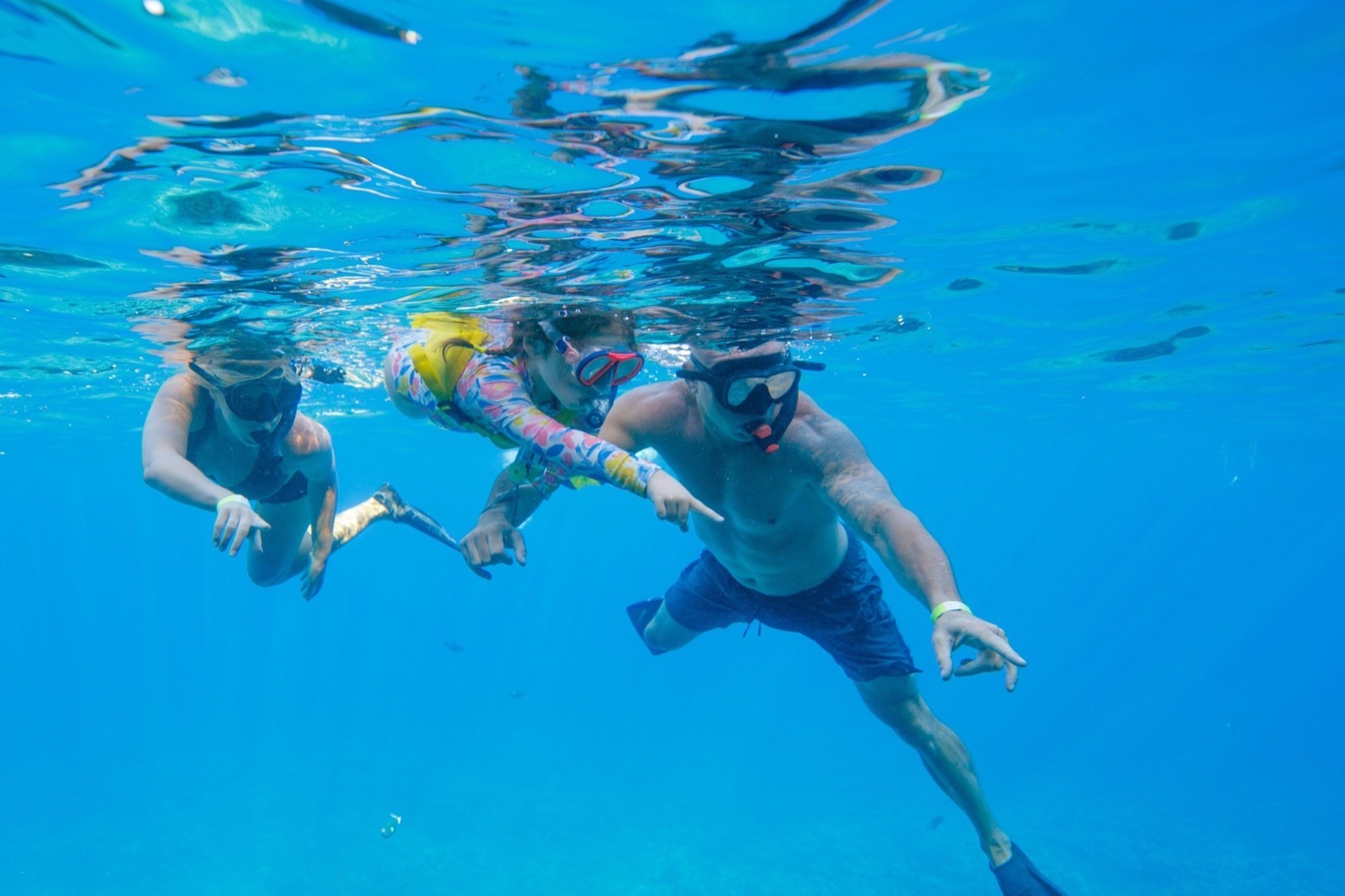 Two adults and a child in a patterned swim shirt and life vest snorkeling together just below the surface