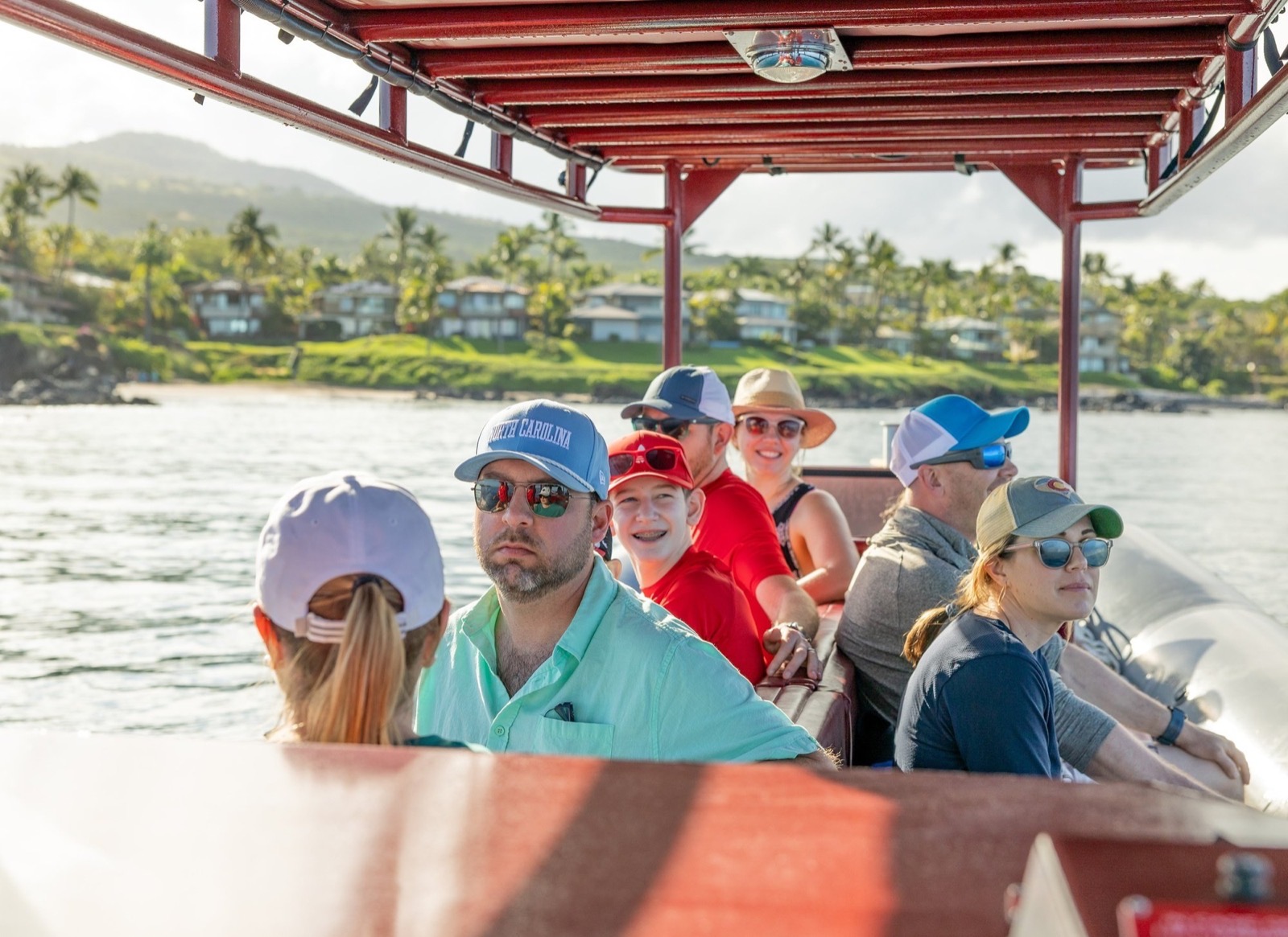 Guests in sunglasses and caps seated on benches under the red canopy of the raft with the palm-lined Kihei shoreline behind them