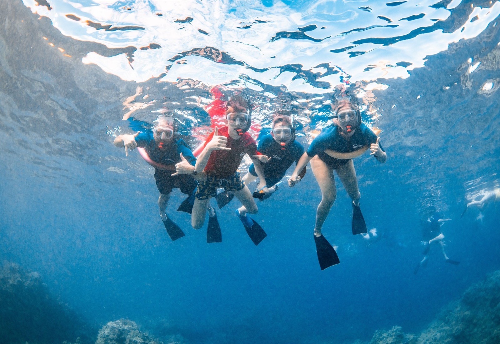 Four snorkelers in masks and fins floating at the surface and flashing shaka signs over a reef
