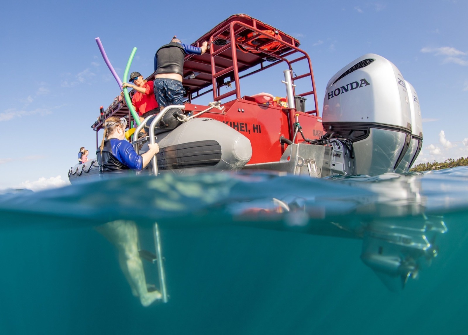 Split view of the red raft from the water, with a guest on the boarding ladder, a crew member holding pool noodles and twin outboard motors