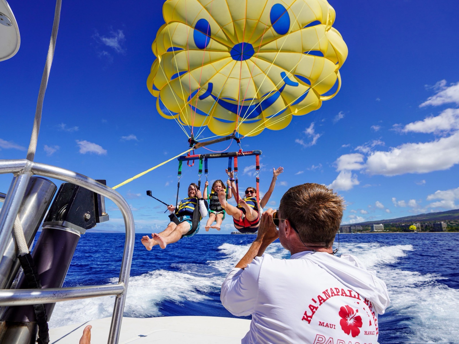 A crew member on the boat deck photographing a family of three in the air under the yellow smiley parasail, with the Kaanapali resorts behind them