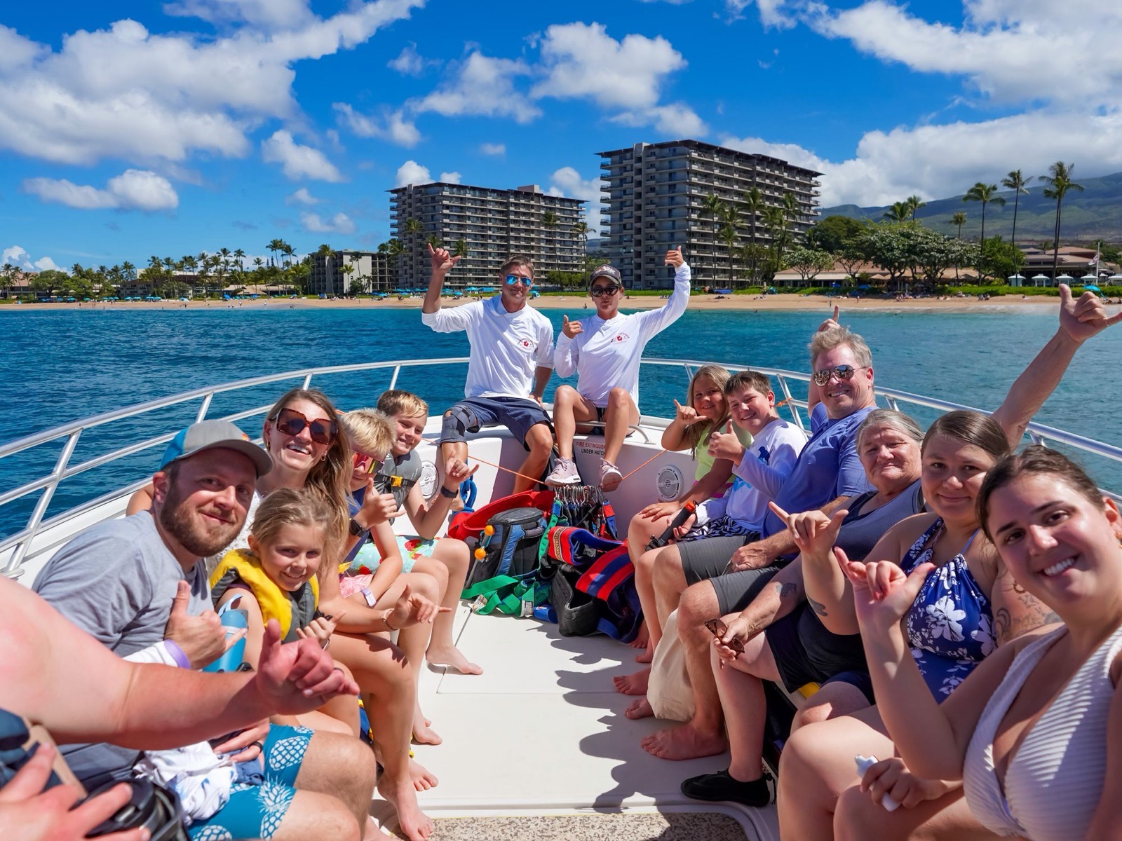 A full boat of smiling guests throwing shakas with two crew members seated at the bow and the Kaanapali Beach resorts and palm trees behind