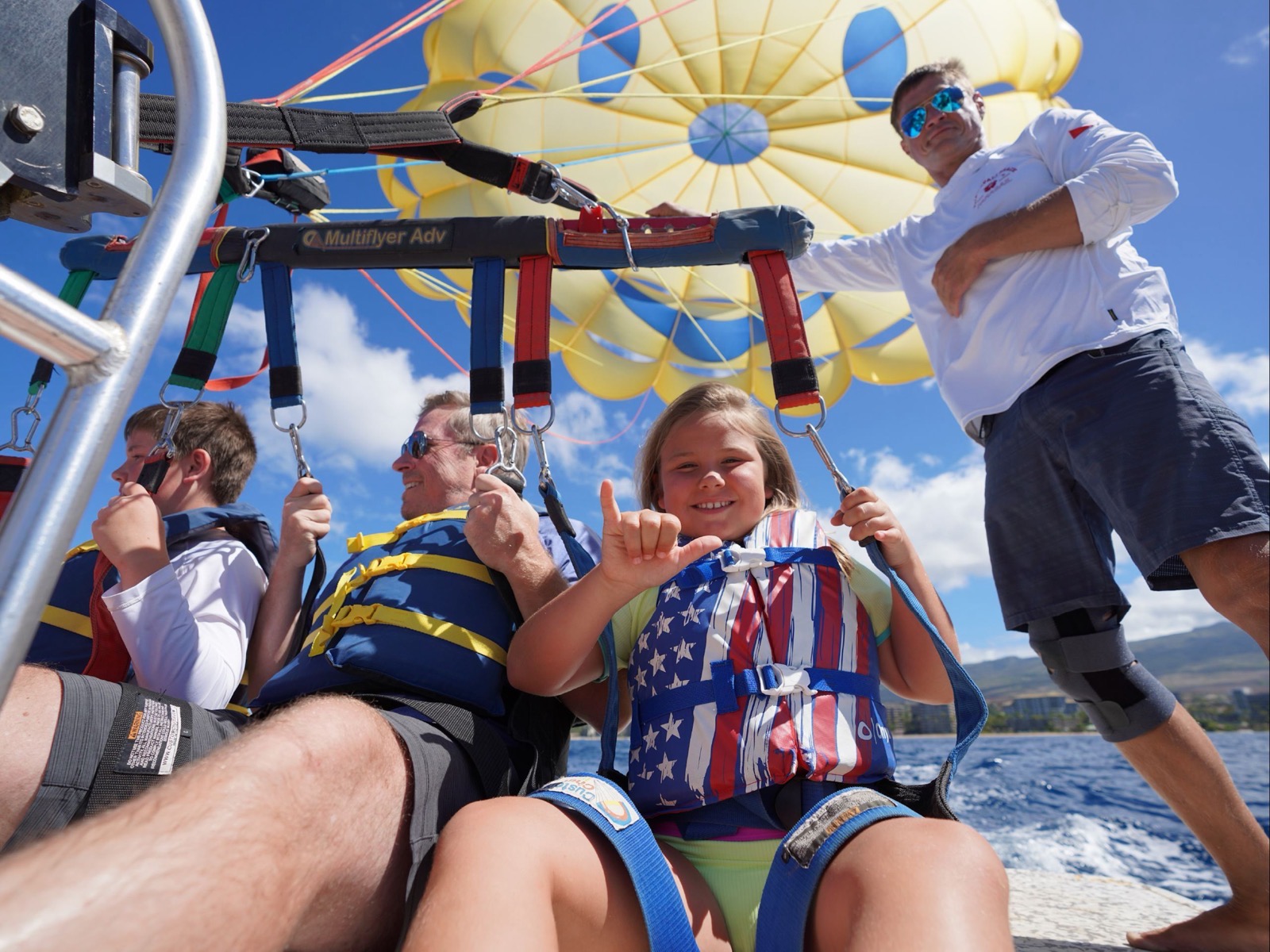 A young flyer in an American-flag life jacket giving a shaka in her harness on the boat deck while a crew member stands beside the parasail canopy