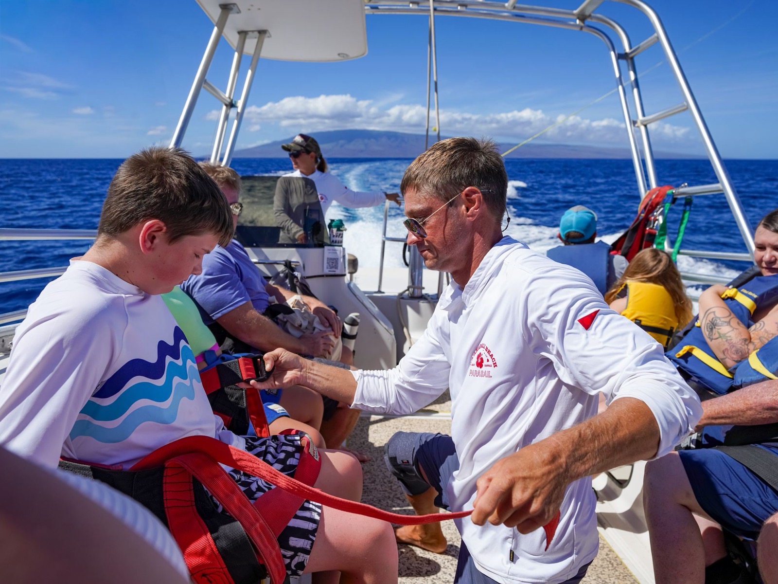A crew member in a white Kaanapali Beach Parasail shirt fitting a harness on a boy seated on the boat, with the island of Lanai on the horizon