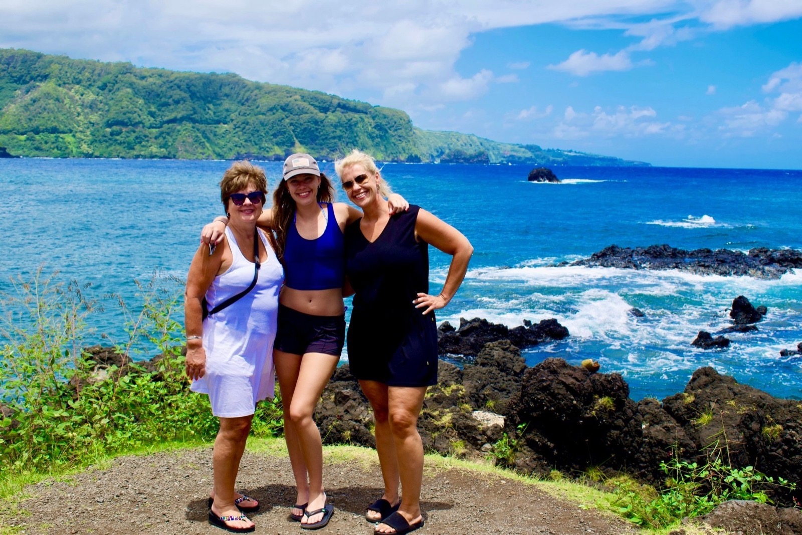 Three women pose arm in arm on a lava-rock overlook above deep blue water with a green headland across the bay