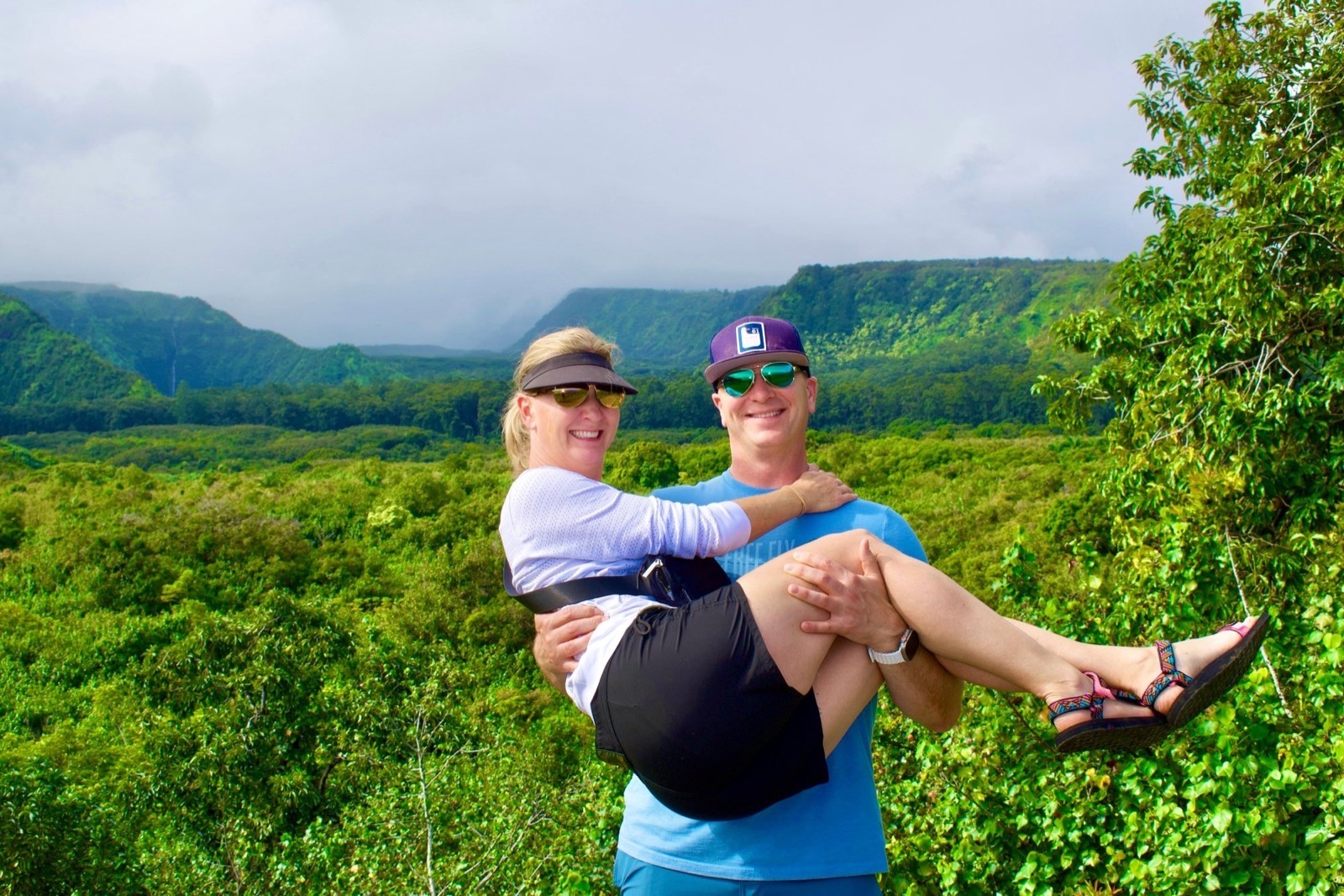 A man in a purple cap lifts a smiling woman in a visor in front of a wide green rainforest valley and cloud-covered ridges