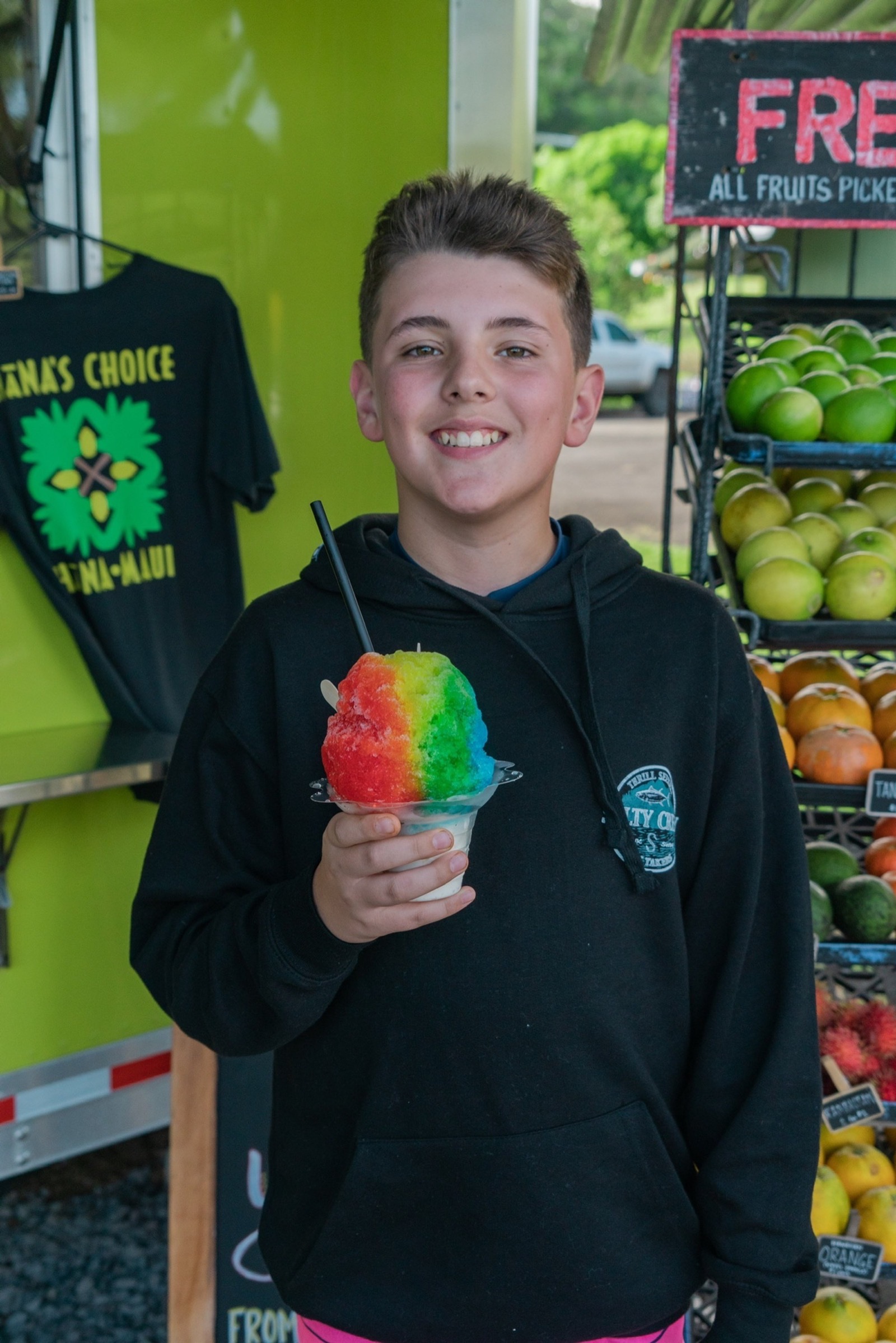A smiling boy holds a rainbow shave ice in front of a lime-green stand with a Hana-Maui shirt and crates of fresh citrus