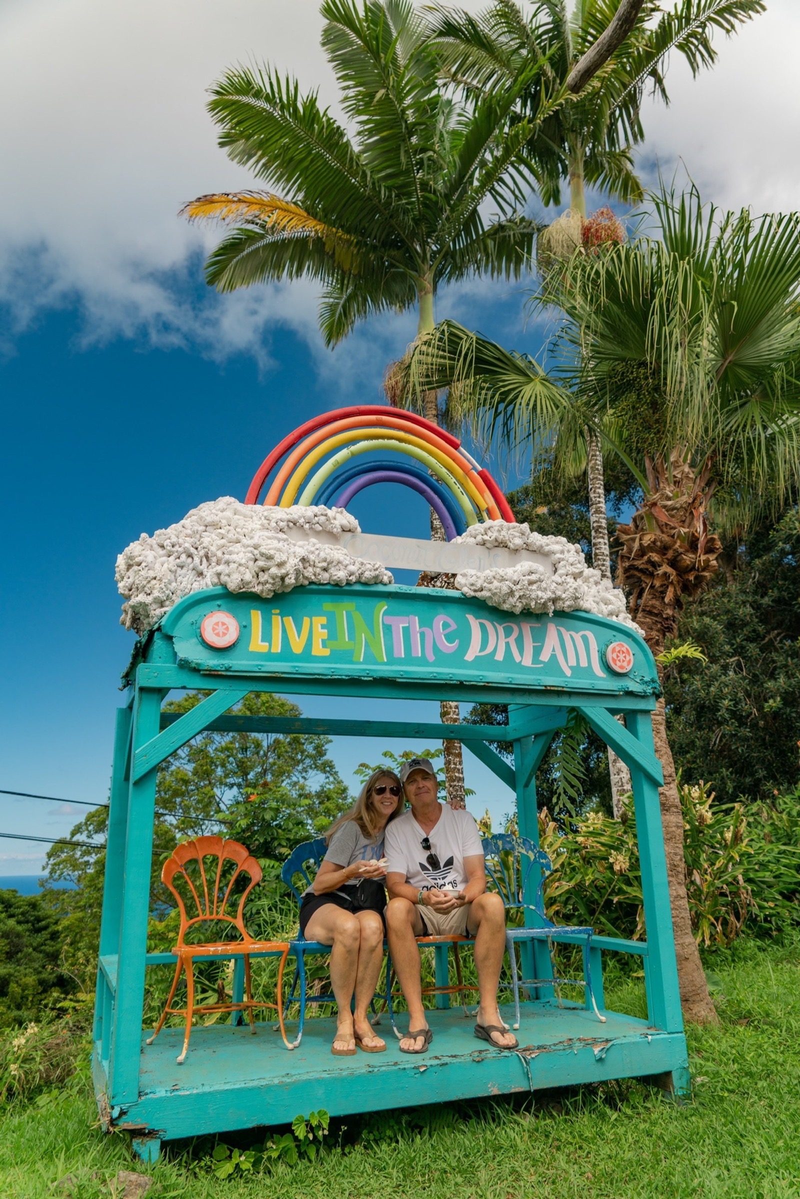 A couple sits on mismatched chairs inside a turquoise roadside shelter topped with a painted rainbow and the words Live in the Dream