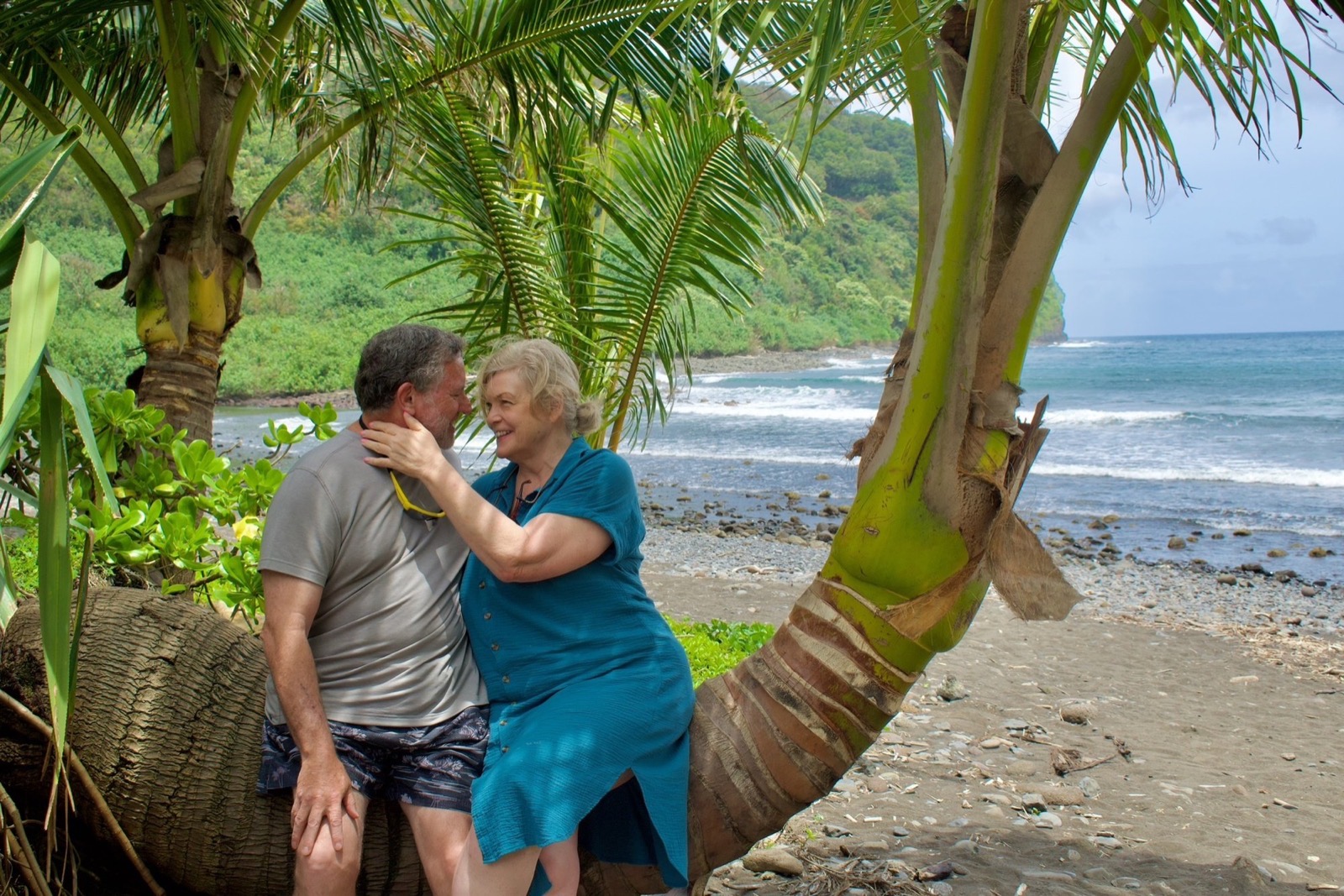 An older couple embraces on a leaning palm trunk beside a pebbly black-sand shoreline with waves rolling in