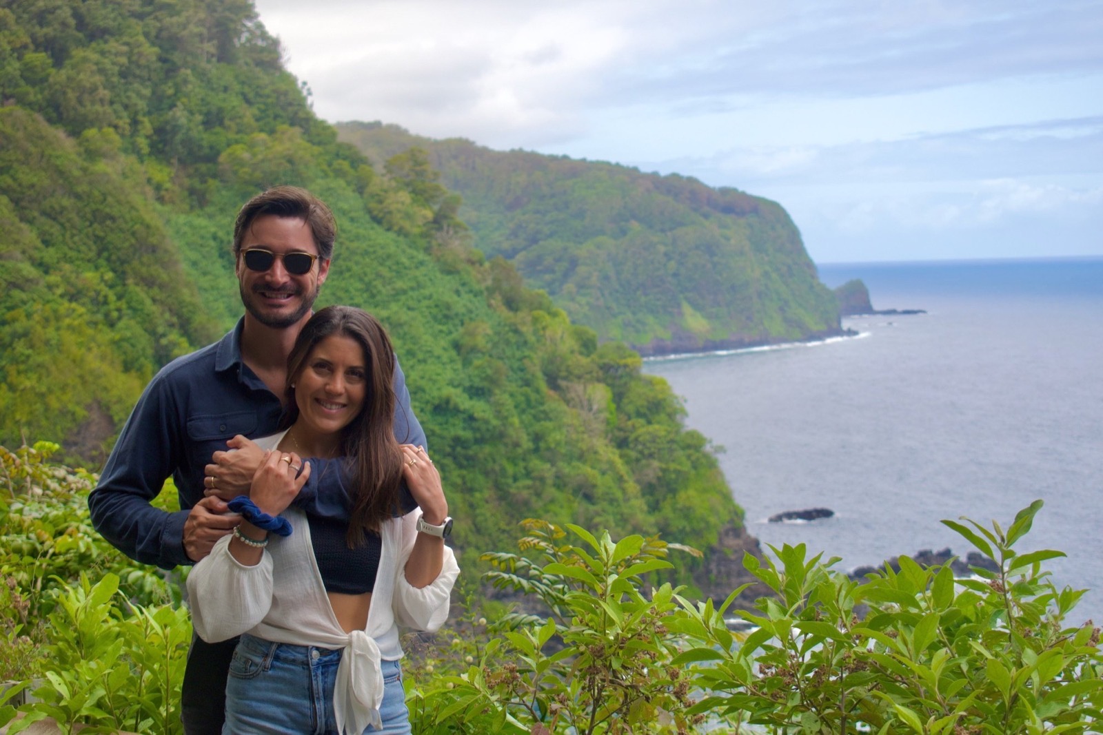 A couple poses on a green coastal cliffside with steep forested headlands dropping to the ocean behind them
