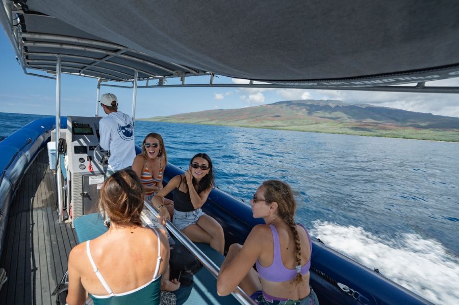 Guests laughing on the seats of the shaded raft while the captain stands at the helm and the coast of Lanai passes behind